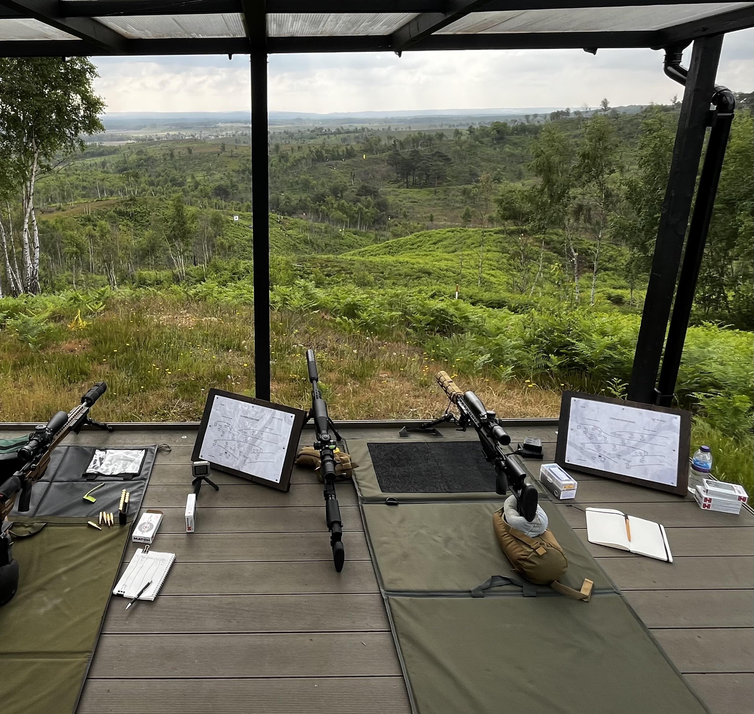 Two precision bolt-action rifles on bipods at a hilltop shooting platform overlooking a long-range target valley