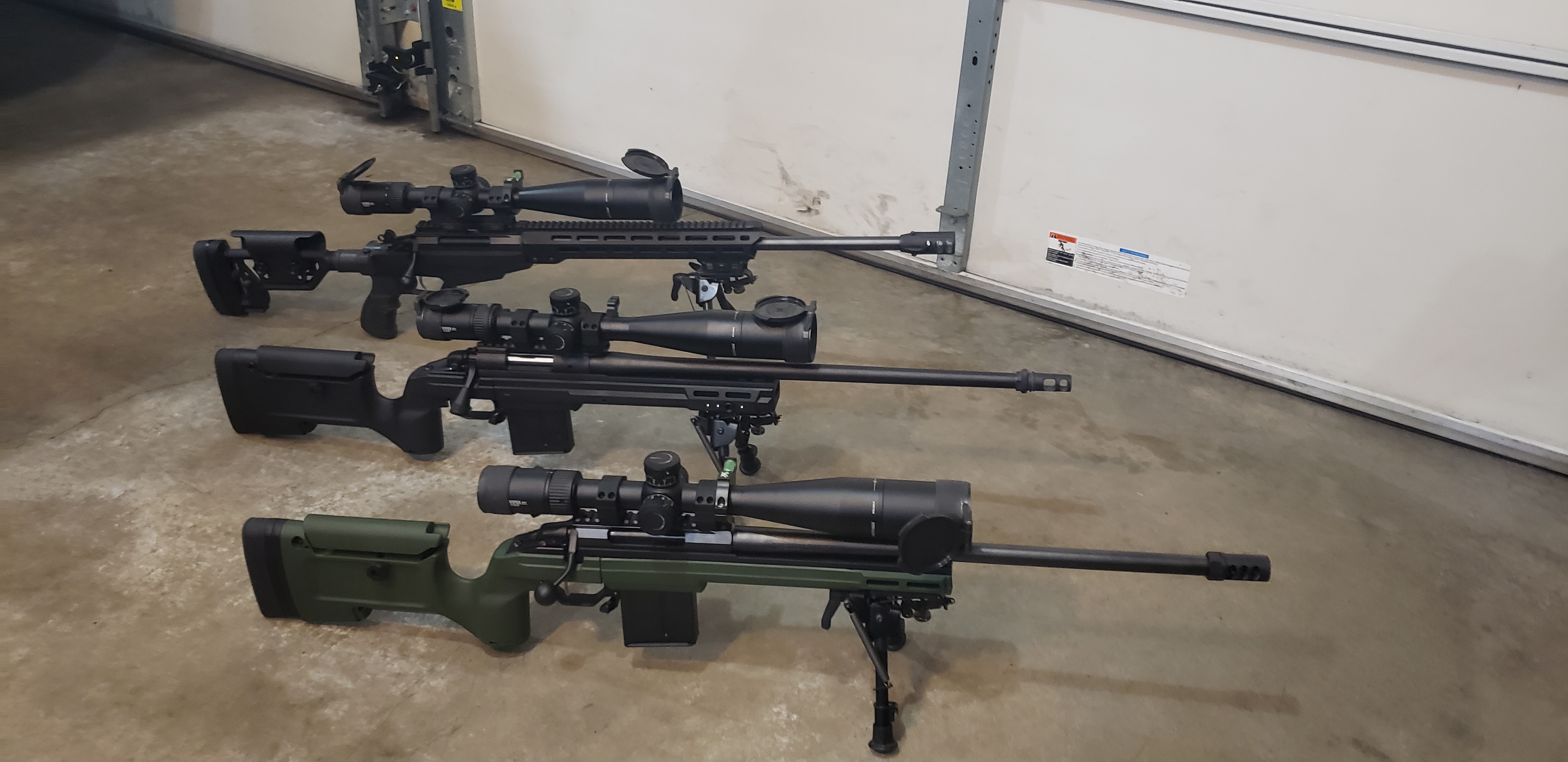 Three scoped precision bolt-action rifles on garage floor, including a green-chassis Tikka T3x TAC A1 on bipod
