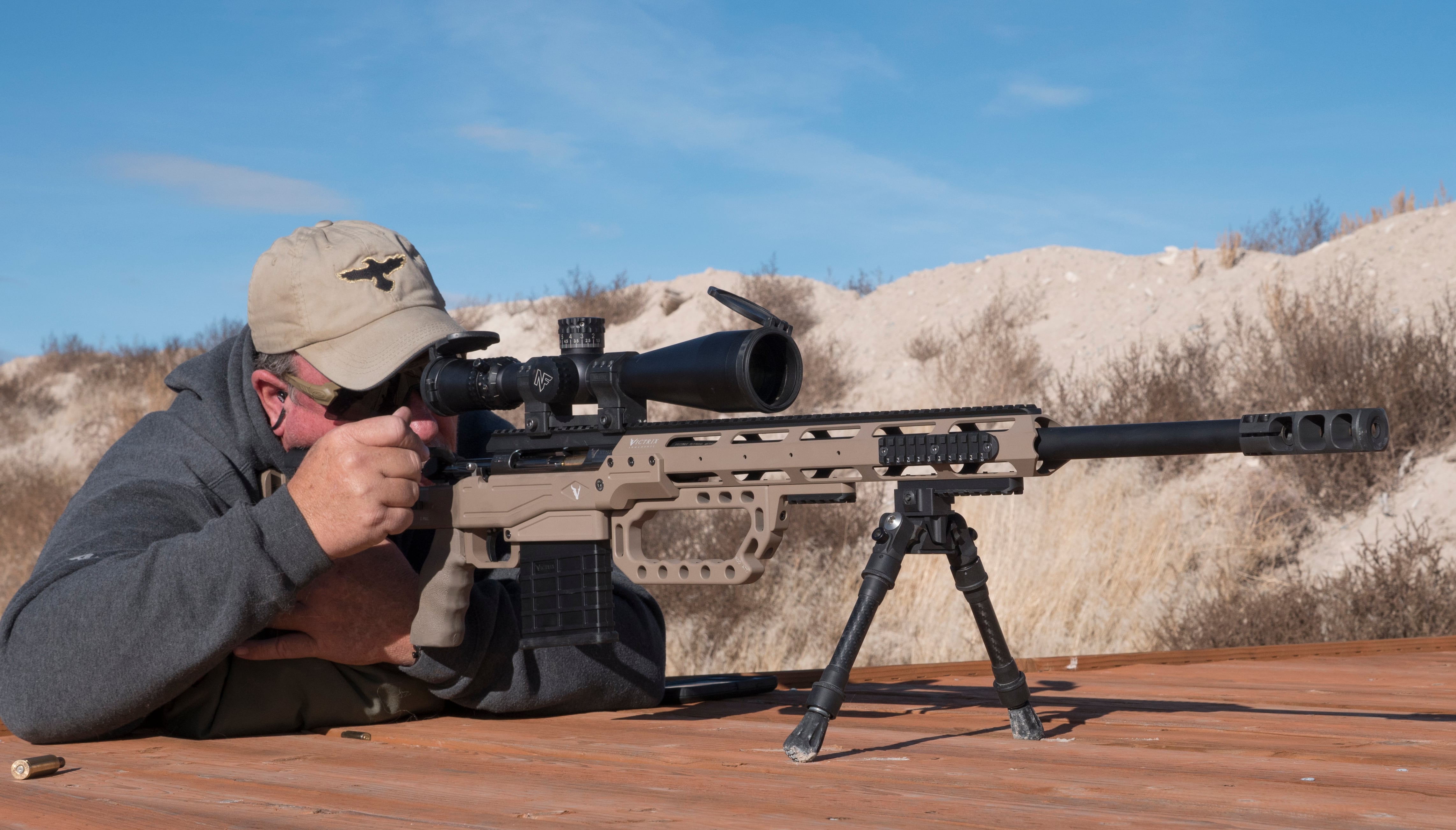 Shooter prone behind a tan chassis precision bolt-action rifle with a Nightforce scope and bipod on a shooting bench at an outdoor range.