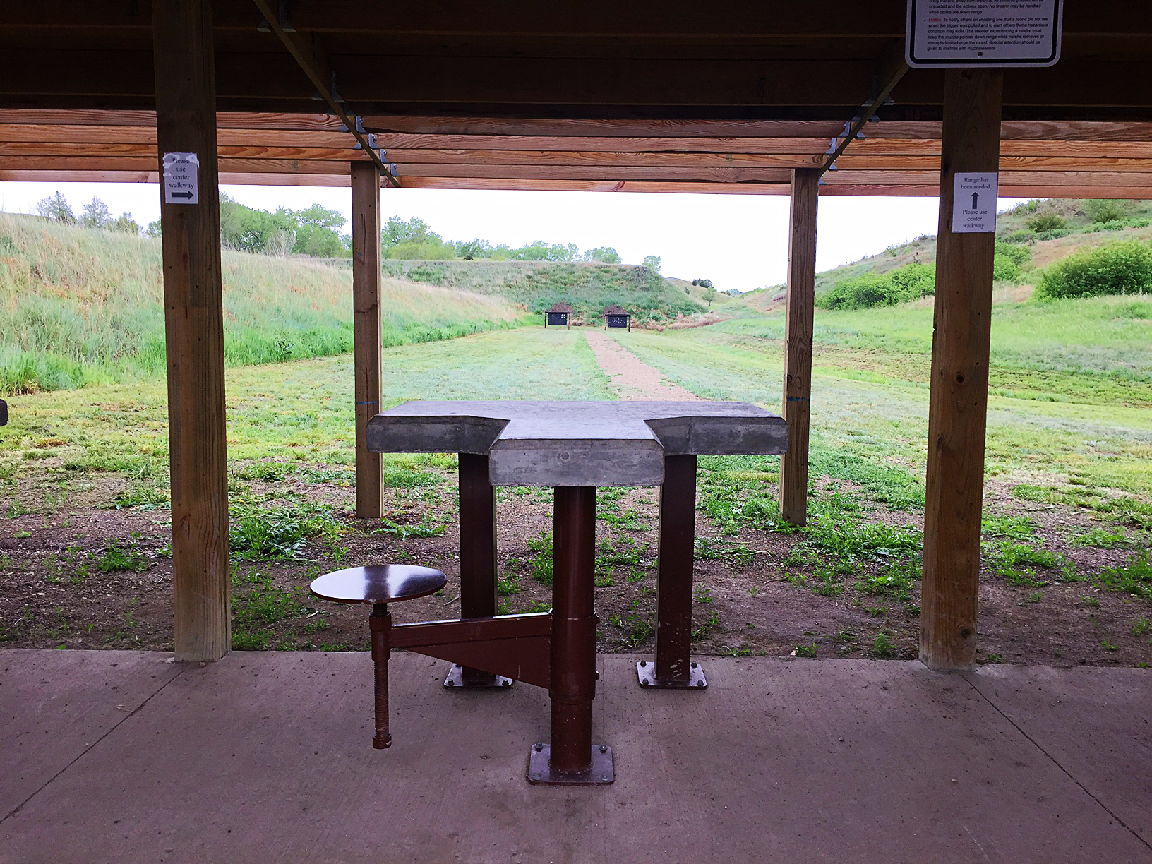 Covered outdoor shooting bench with concrete rest and metal seat, looking downrange toward target frames set against a grass berm.