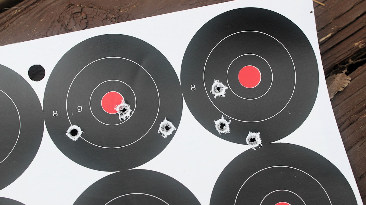 Paper shooting target with multiple bullet holes clustered near the center rings of two circular aiming points.