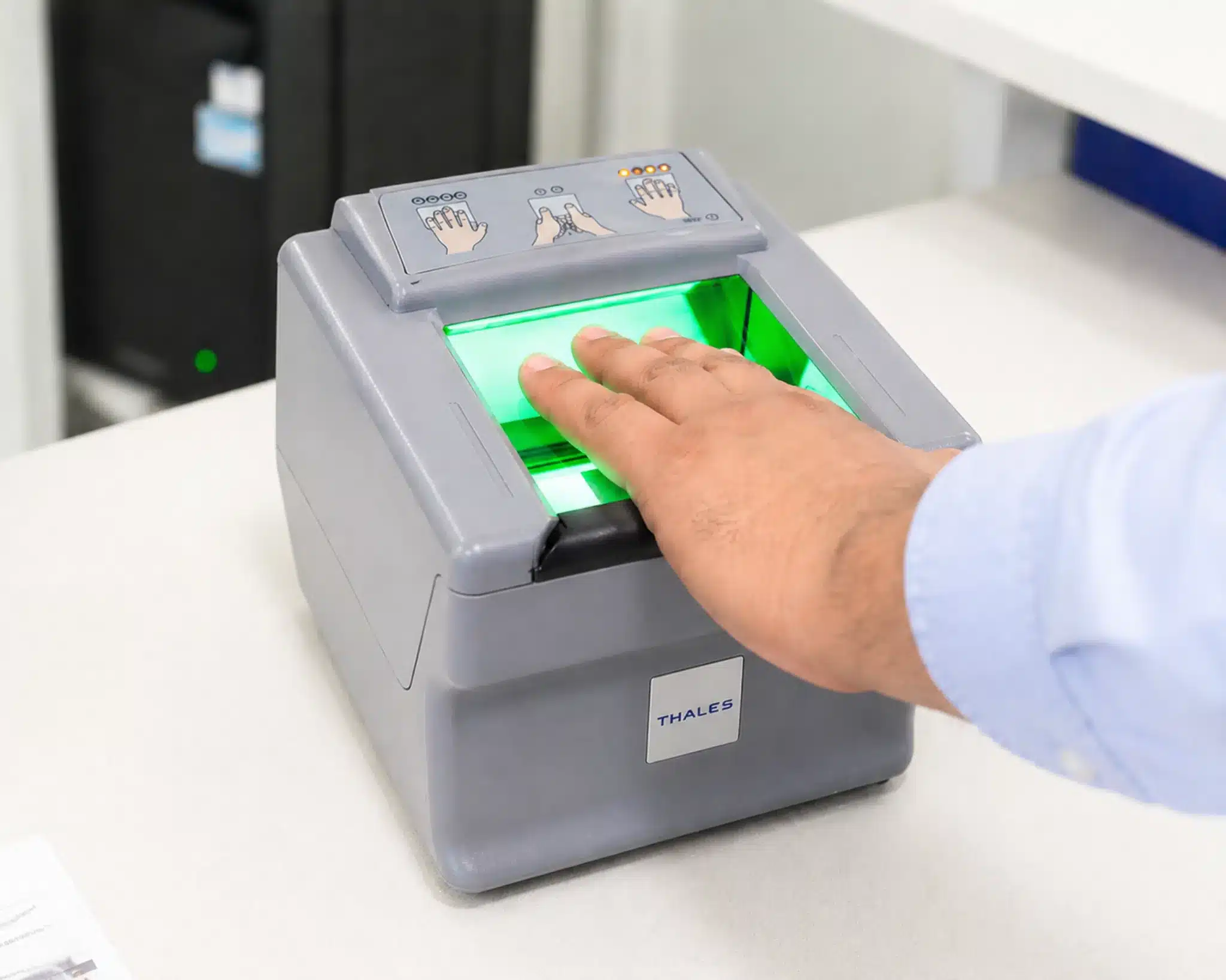 A person pressing four fingers onto the illuminated green scanning surface of a Thales live scan fingerprint machine on a white desk.