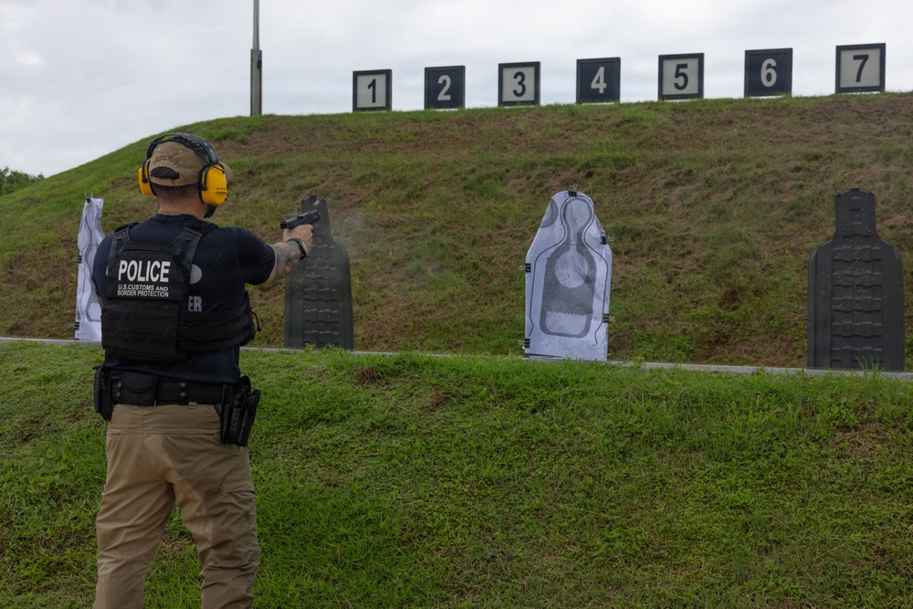 A U.S. Customs and Border Protection officer in a police vest fires a handgun at numbered silhouette targets on an outdoor shooting range.