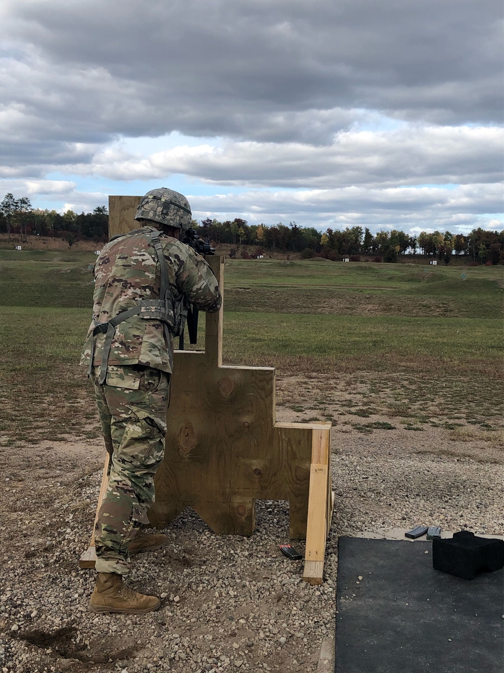 Army soldier in OCP uniform firing from behind a wooden barricade on an outdoor qualification range.