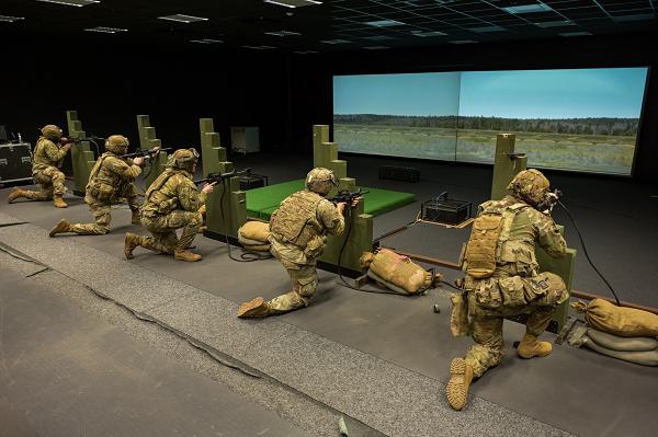 Soldiers in camouflage kneel with rifles aimed at a projected outdoor scene on an EST simulation range.