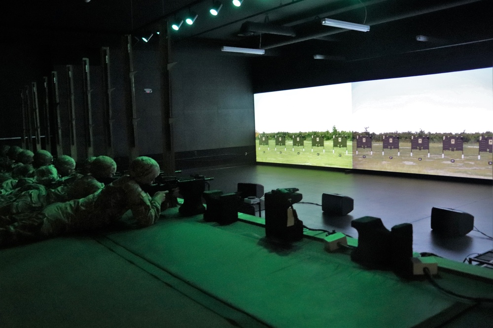 Soldiers prone with rifles in a darkened indoor marksmanship simulator, aiming at a large projected target screen.