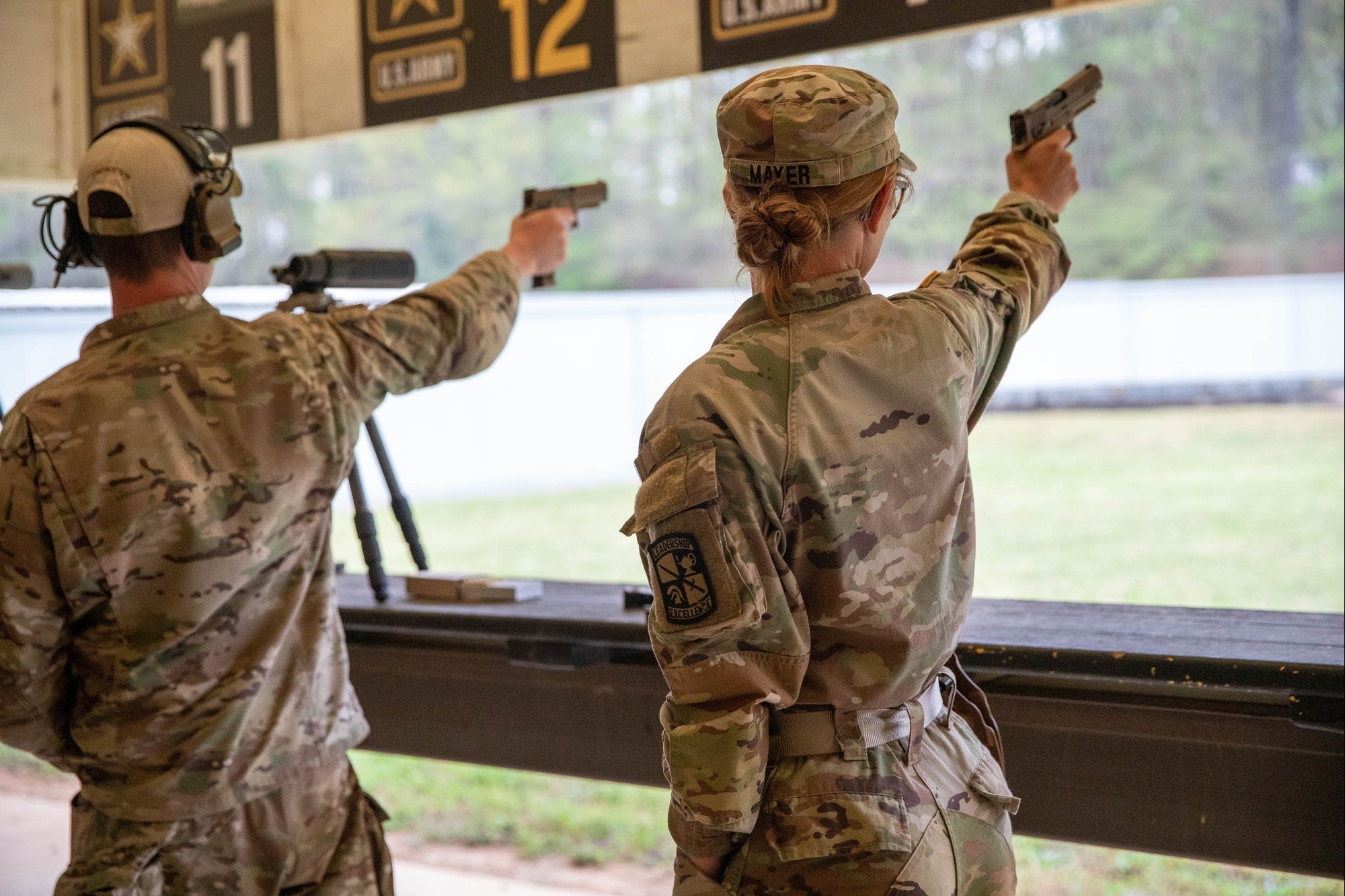 Two U.S. Army soldiers in OCP uniforms firing pistols at a numbered indoor shooting range.
