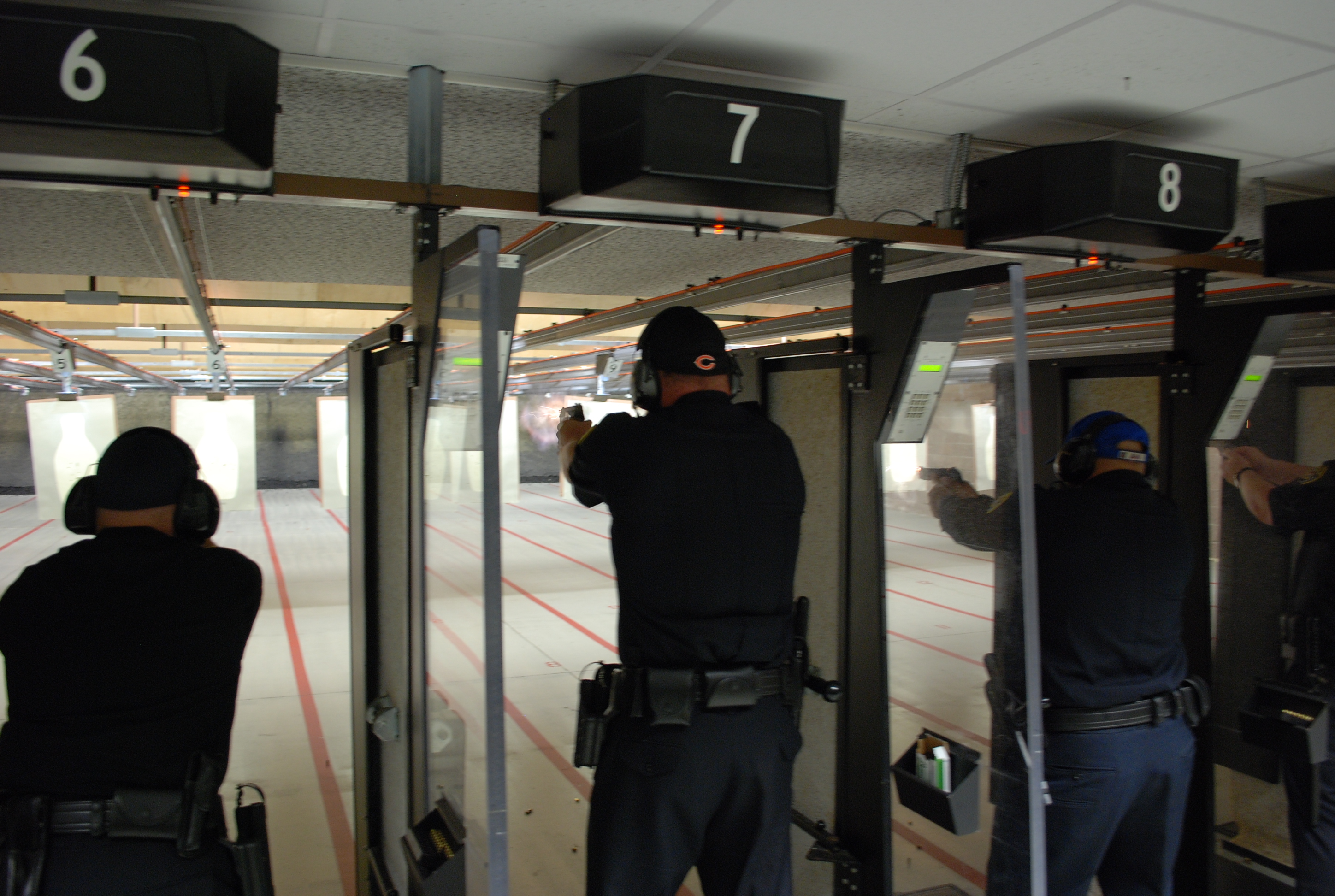 Three shooters in black clothing fire handguns from numbered lanes 6, 7, and 8 at an indoor shooting range.
