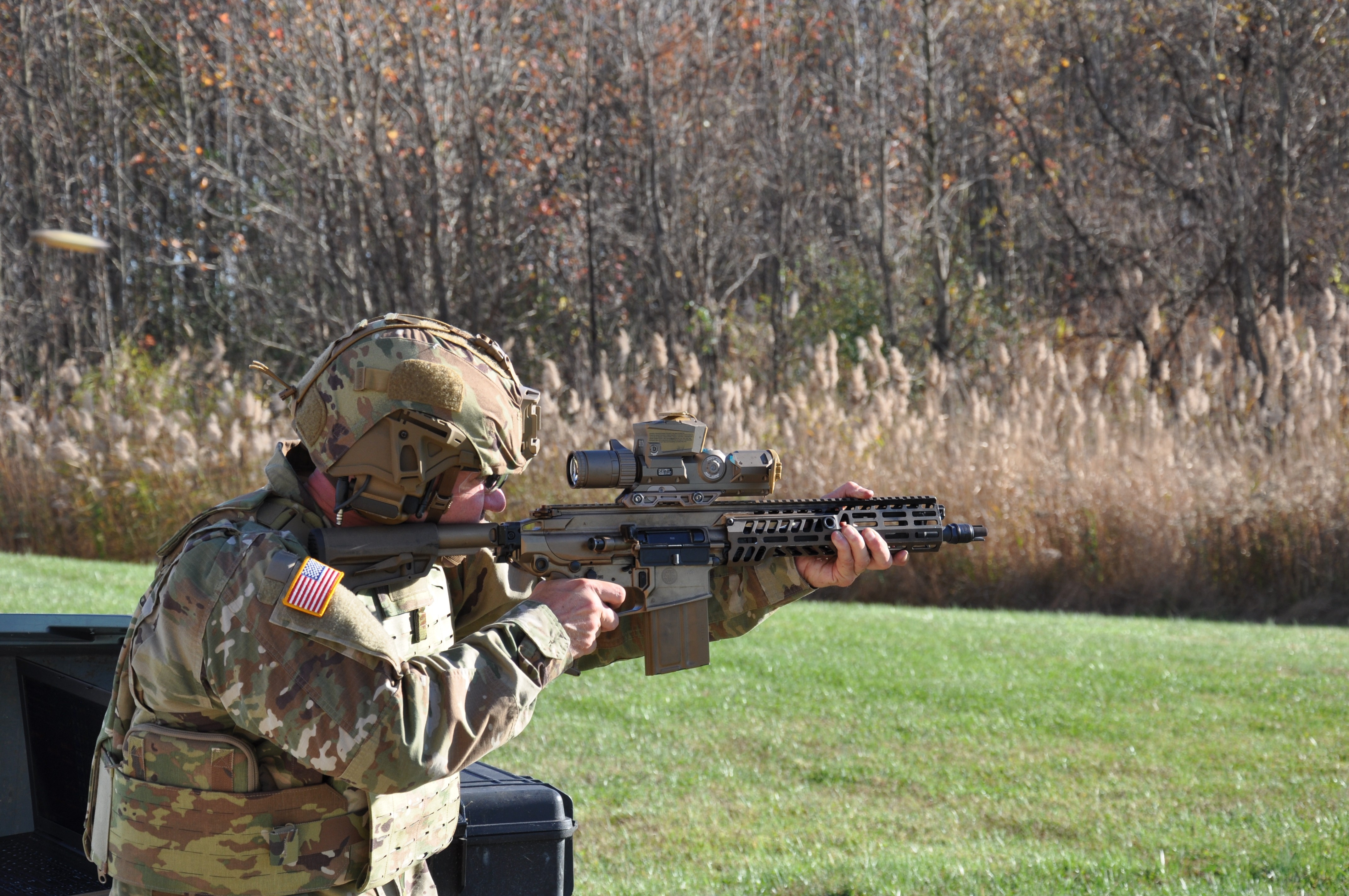 U.S. Army soldier in OCP uniform aiming a next-generation carbine with optic at an outdoor firing range.