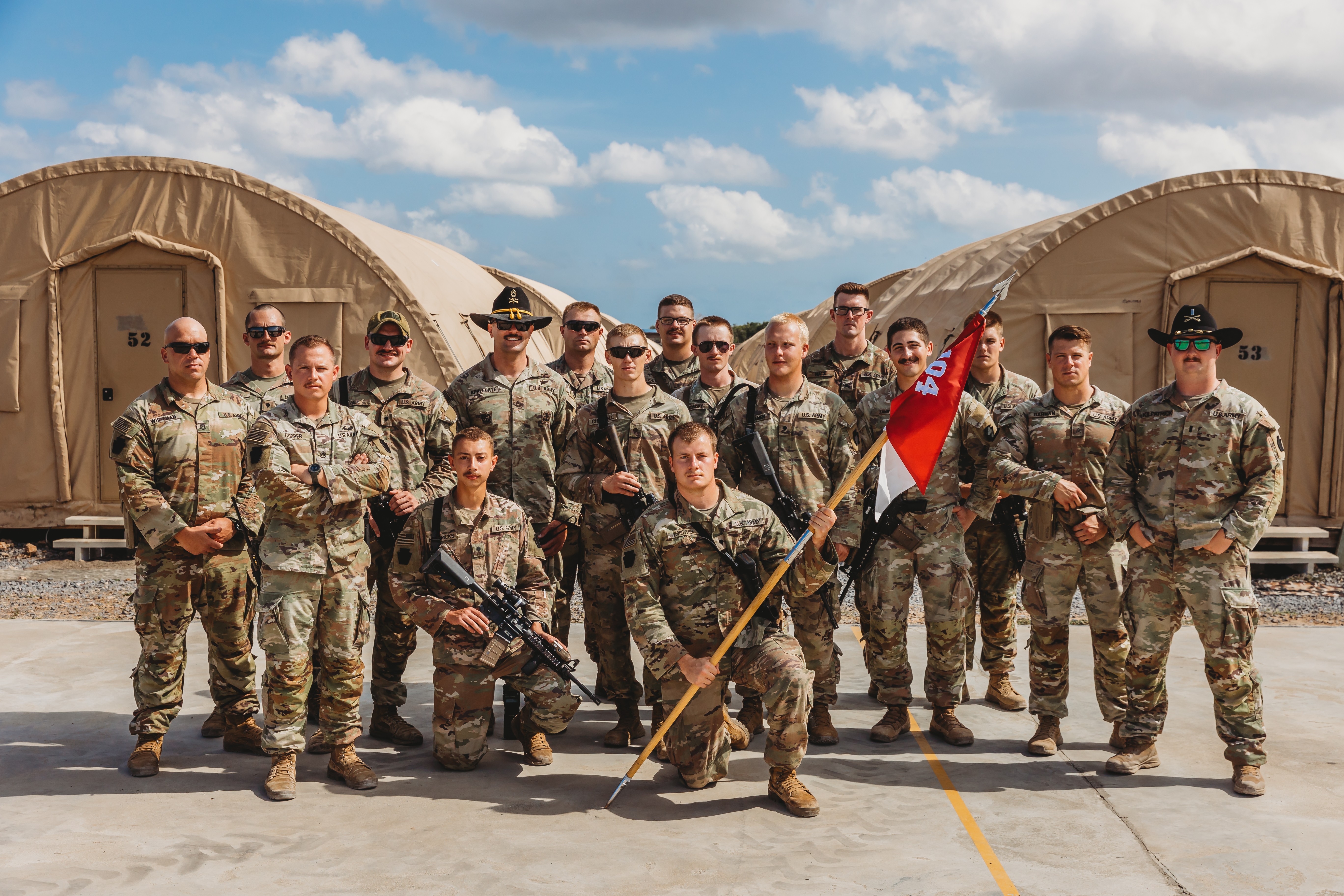 Group of U.S. Army National Guard soldiers in OCP uniforms posing in front of military tents with rifles and a unit guidon.