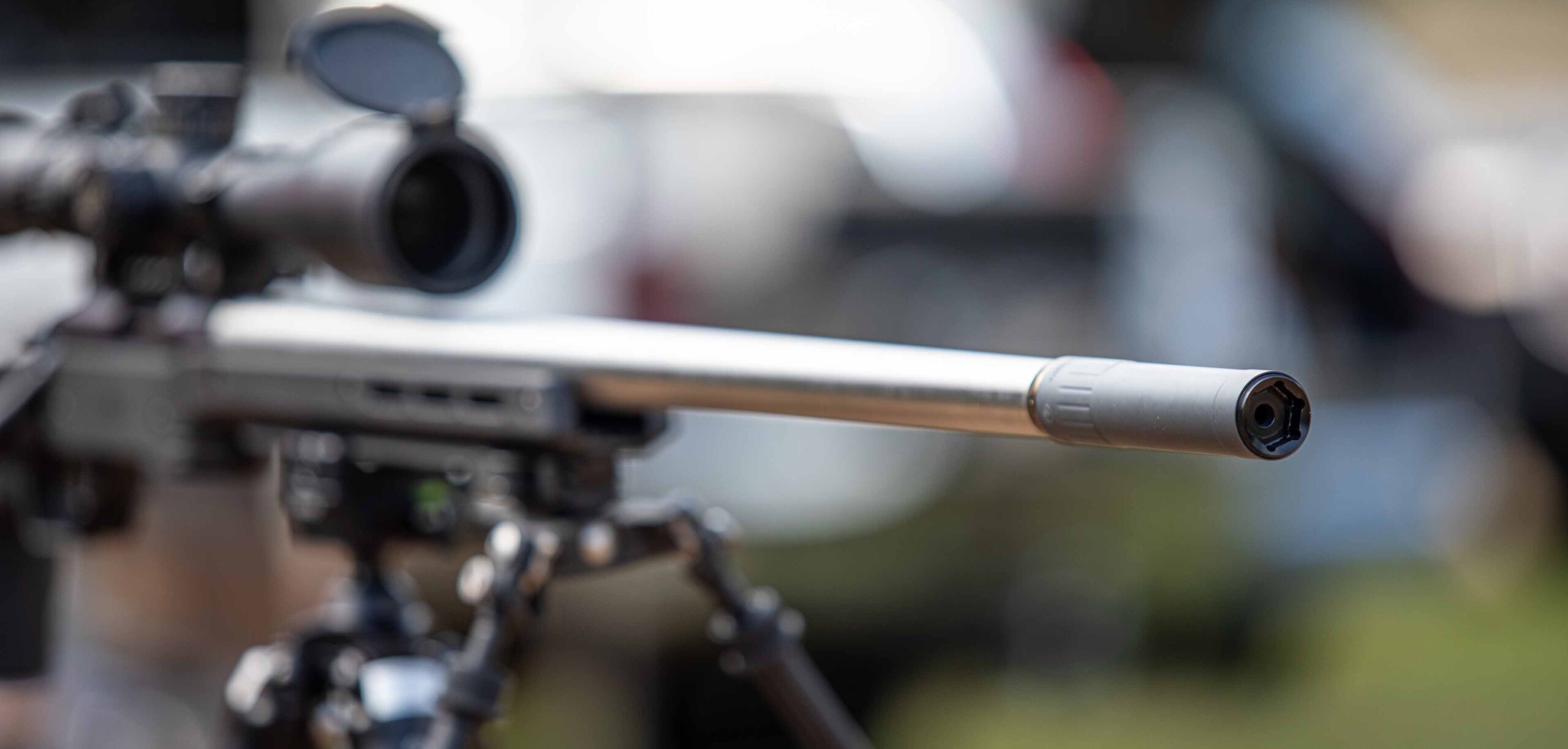 Close-up of a bolt-action rifle barrel with a muzzle device, bipod legs and riflescope visible in the background.