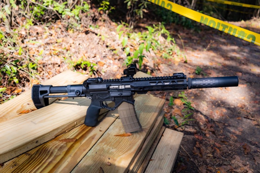 AR-style pistol with suppressor, red dot optic, pistol brace, and FDE magazine resting on a wooden bench at an outdoor range.