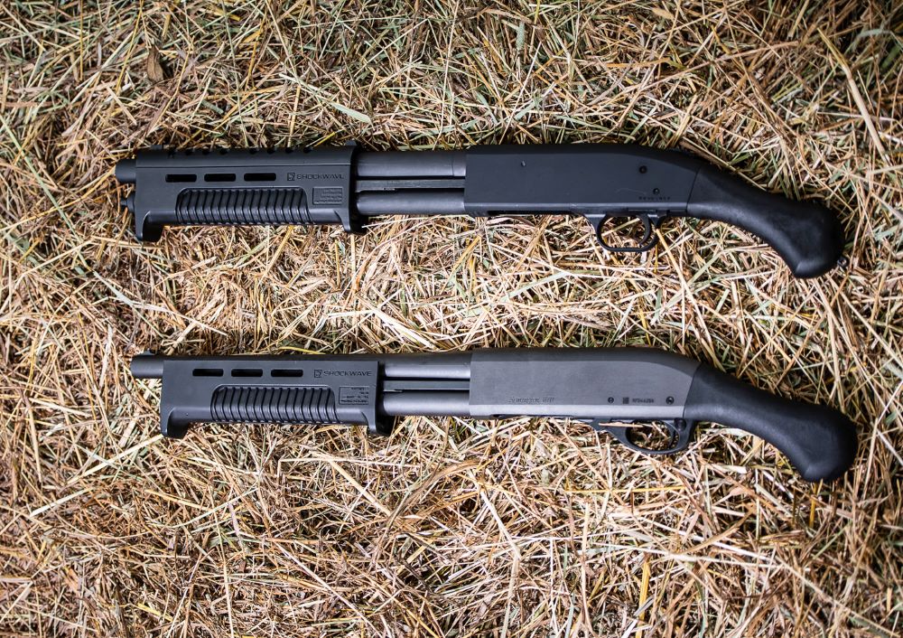 Two black Mossberg Shockwave short-barreled pump-action firearms with bird's-head pistol grips lying on dry grass, viewed from above.