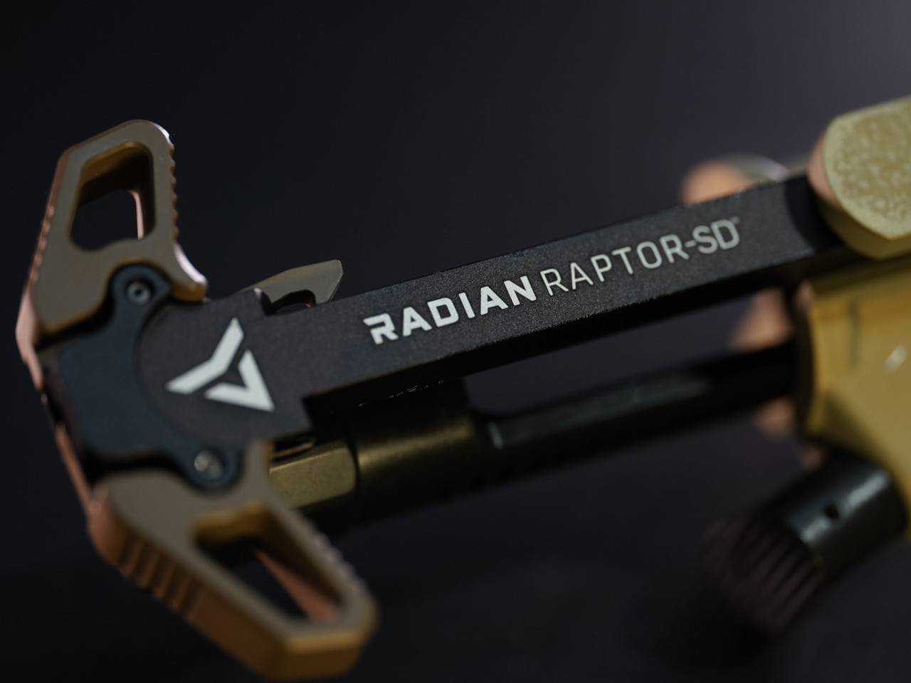 Close-up of a Radian Raptor-SD charging handle in black and bronze finish against a dark background, showing the Radian logo and model name engraved on the body.