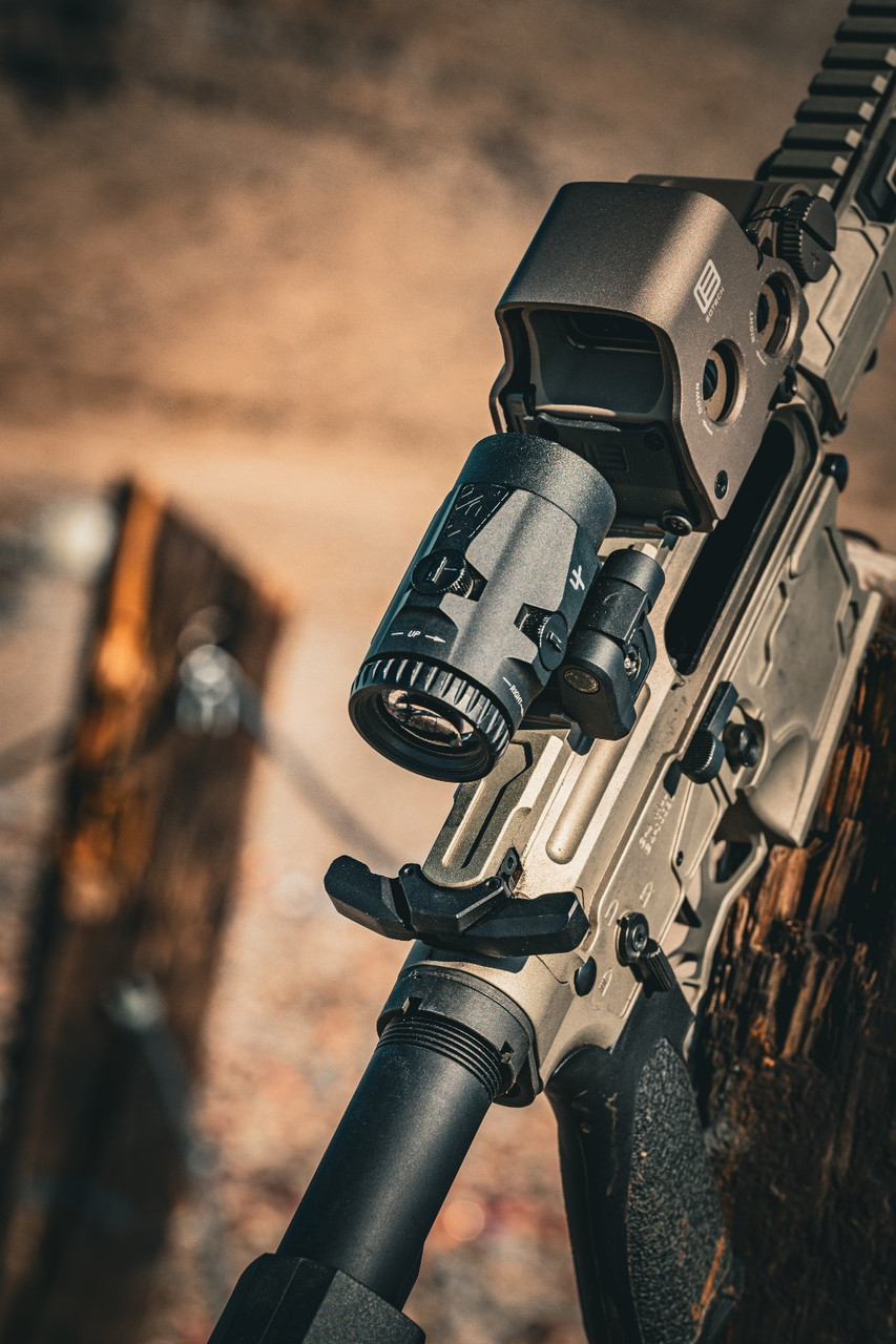Close-up of an AR-15 upper receiver with an EOTech holographic sight and magnifier mounted on a Picatinny rail, leaned against a wooden post outdoors.