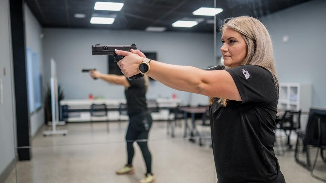 Two women in a classroom or training facility extending semi-automatic pistols in a shooting stance, practicing pistol presentation drills.