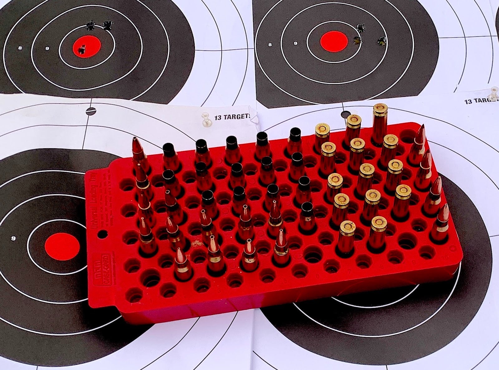 Red reloading tray filled with rifle cartridges at various stages of completion, placed on top of paper shooting targets with bullet holes.