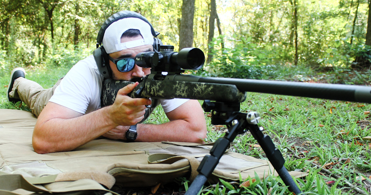 Shooter prone behind a scoped bolt-action rifle on a bipod, aiming downrange in an outdoor wooded setting.