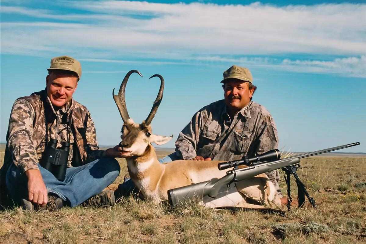Two hunters kneeling behind a harvested pronghorn antelope on open grassland, with a scoped bolt-action rifle on a bipod displayed in the foreground.