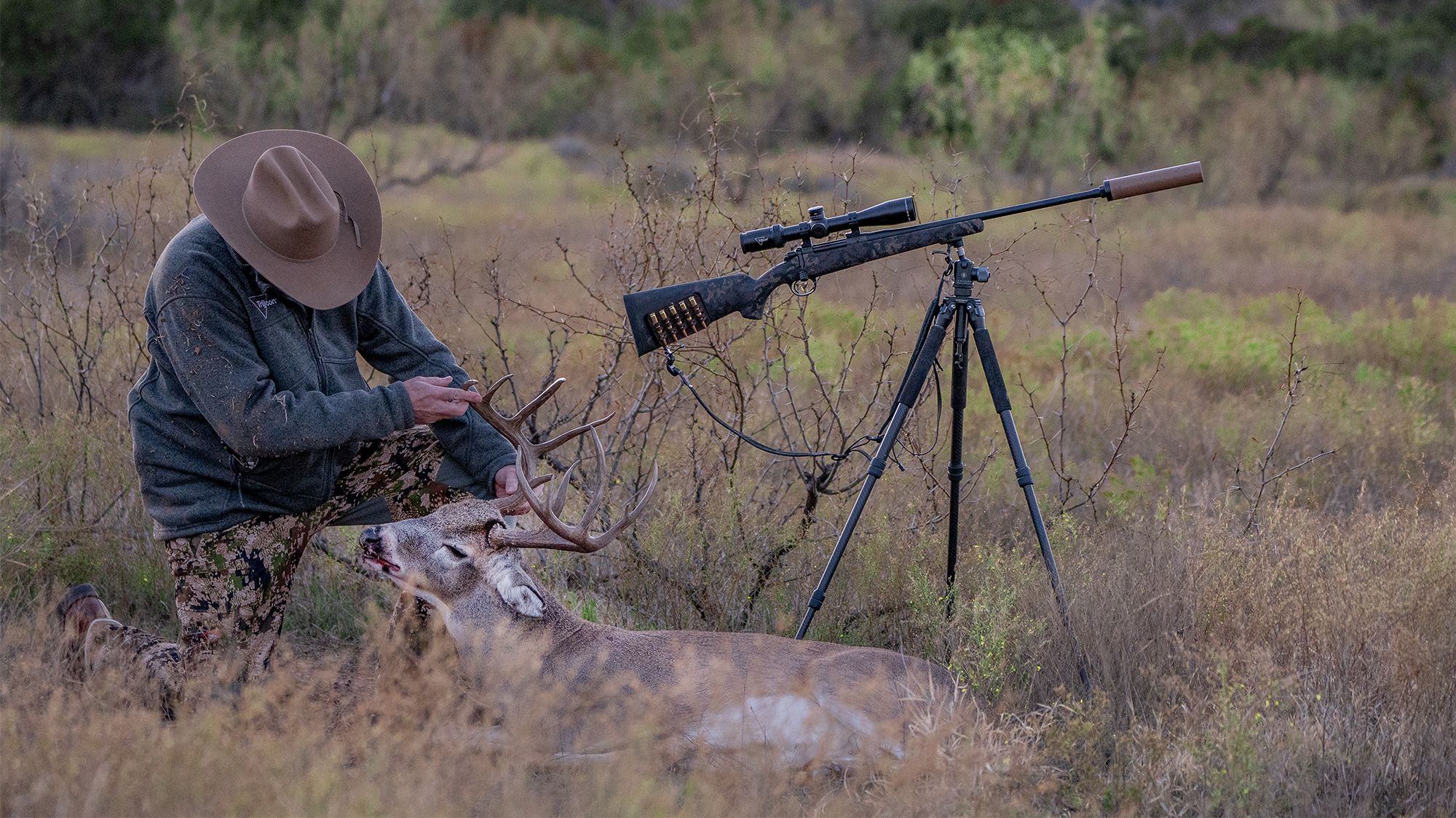 Hunter kneeling next to harvested whitetail buck with suppressed bolt-action rifle on tripod in brushy field.