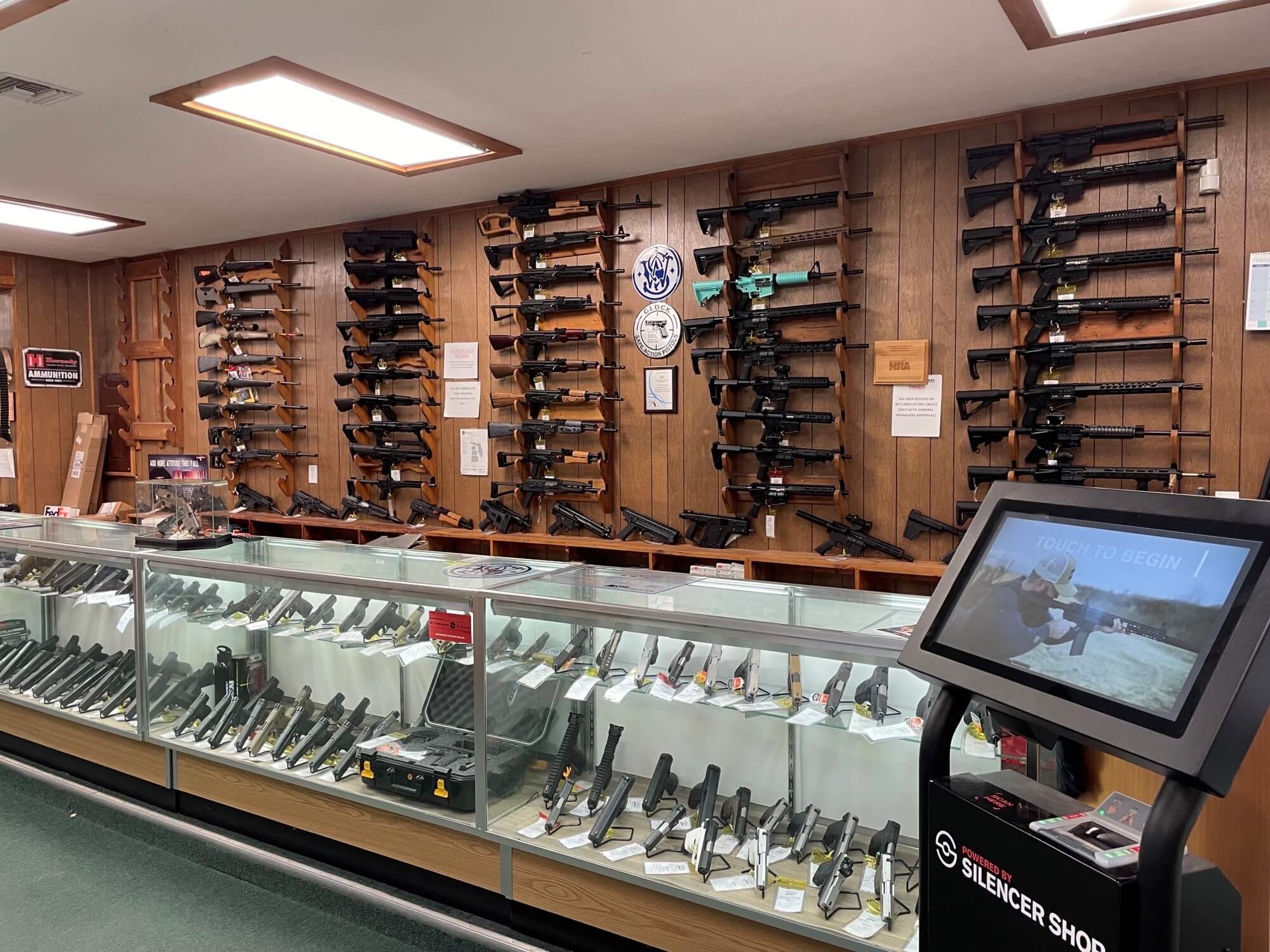 Gun store interior with AR rifles on wall, handguns in glass case, and a Silencer Shop kiosk in foreground.