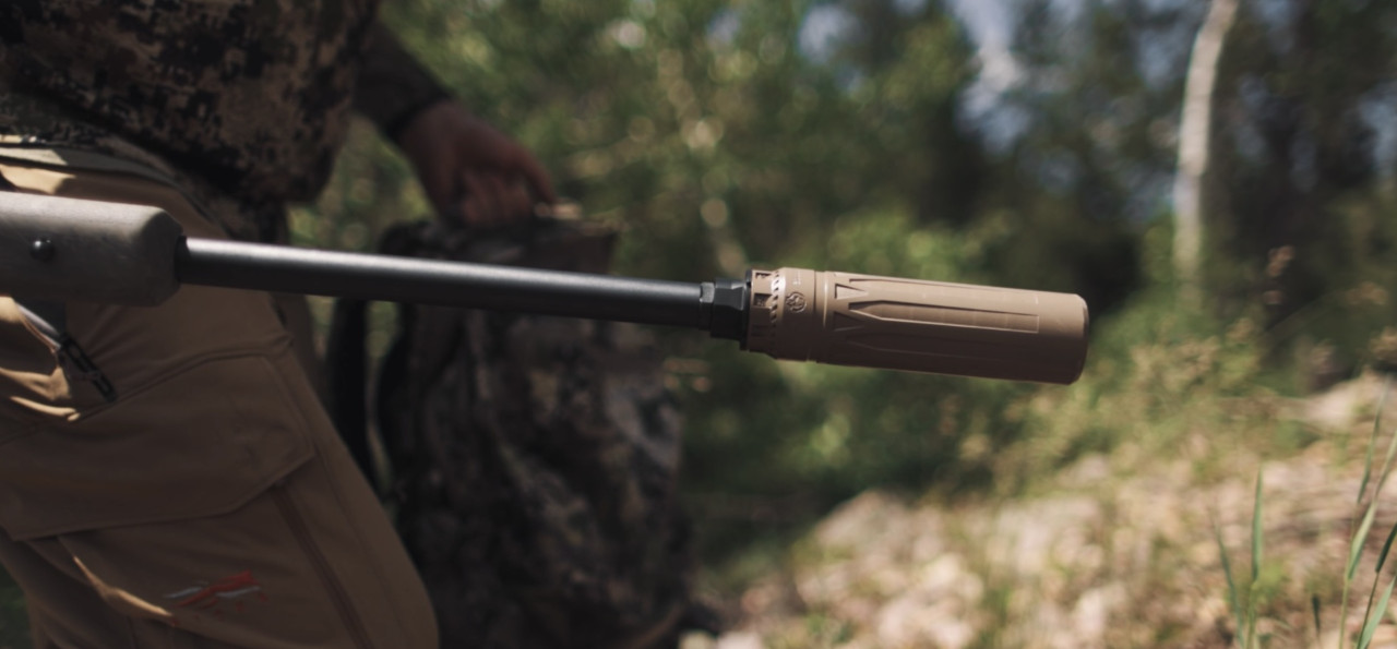 Hunter holding bolt-action rifle with tan suppressor attached, outdoors in natural light.