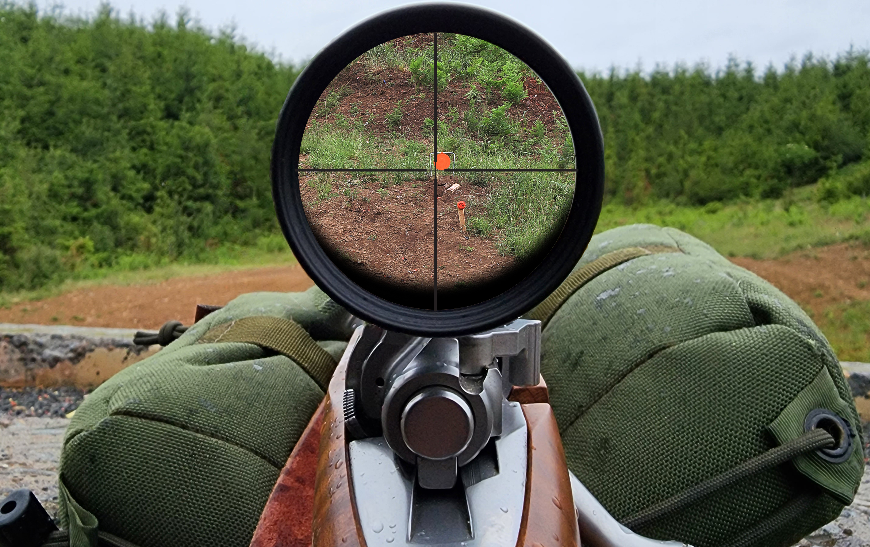 Rear view of a scoped bolt-action rifle on a shooting bag, with a clear scope image showing crosshairs on an orange target at an outdoor range.