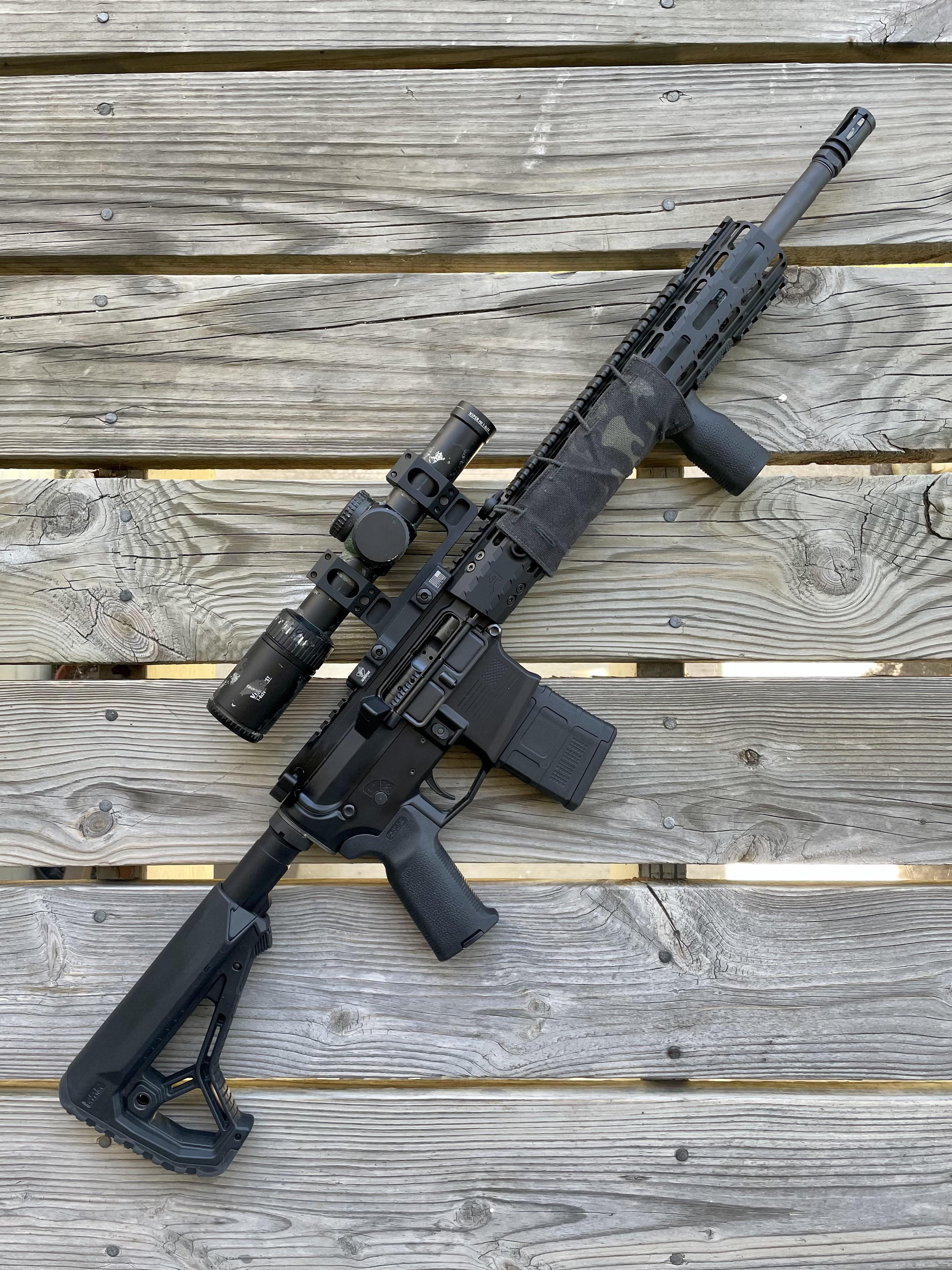 AR-style rifle with LPVO scope mounted on a tall mount, laid flat on a wooden surface, with a folding/adjustable stock and M-LOK handguard.