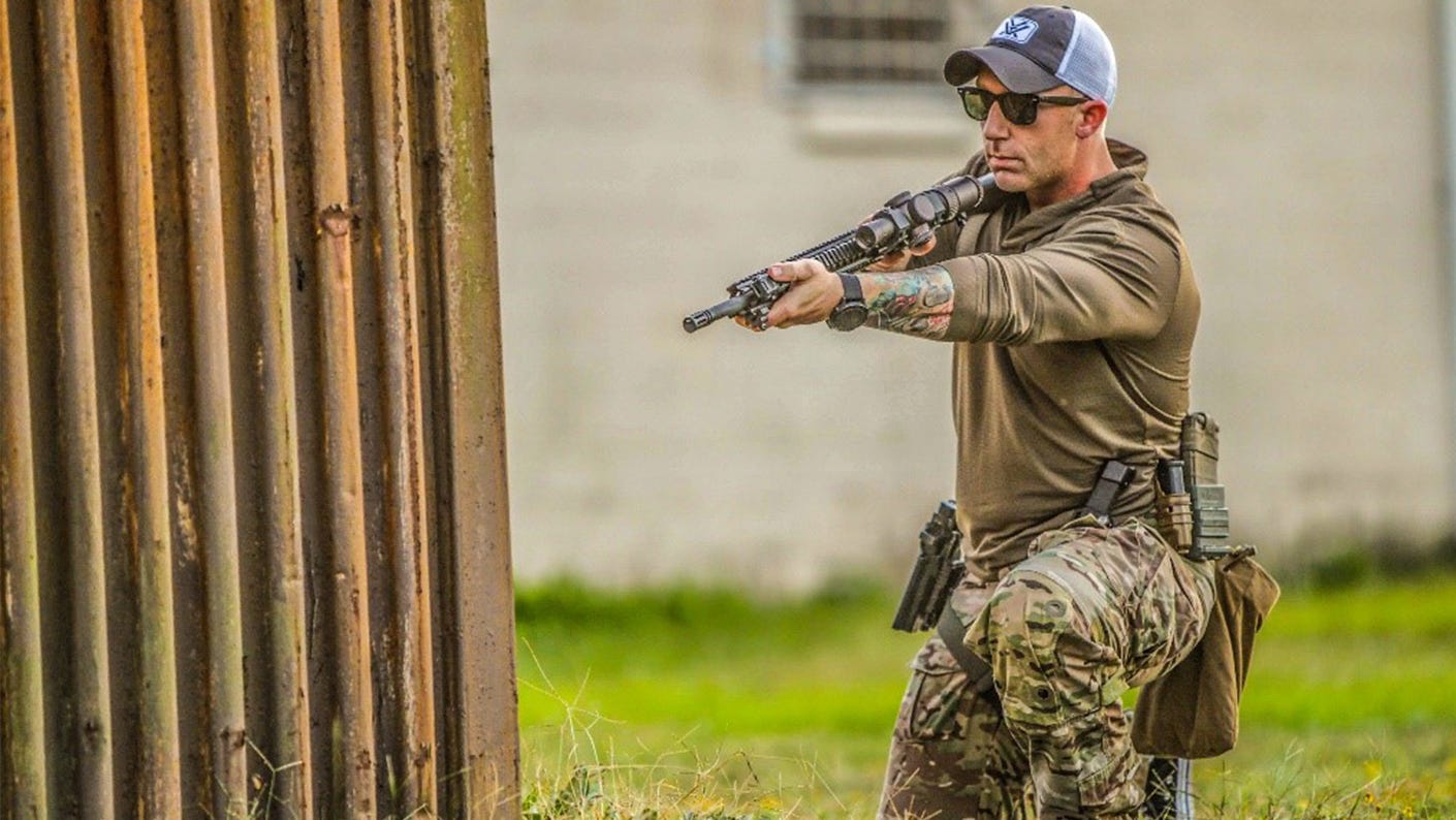 Shooter in kneeling position aiming a scoped AR-style rifle around a metal barrier, with an LPVO mounted on the rifle.