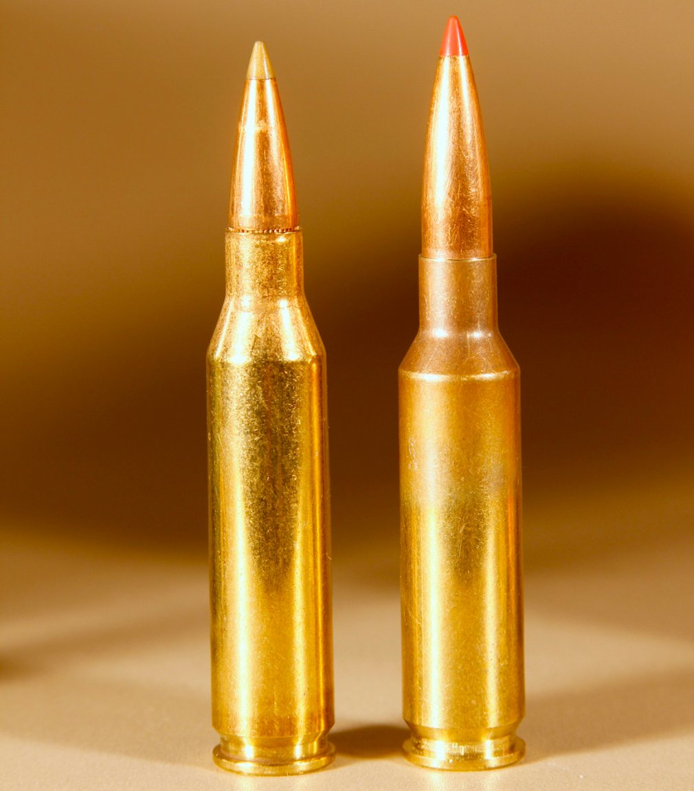 Two rifle cartridges standing upright side by side: a longer brass cartridge on the left and a slightly shorter cartridge with a red polymer tip on the right.