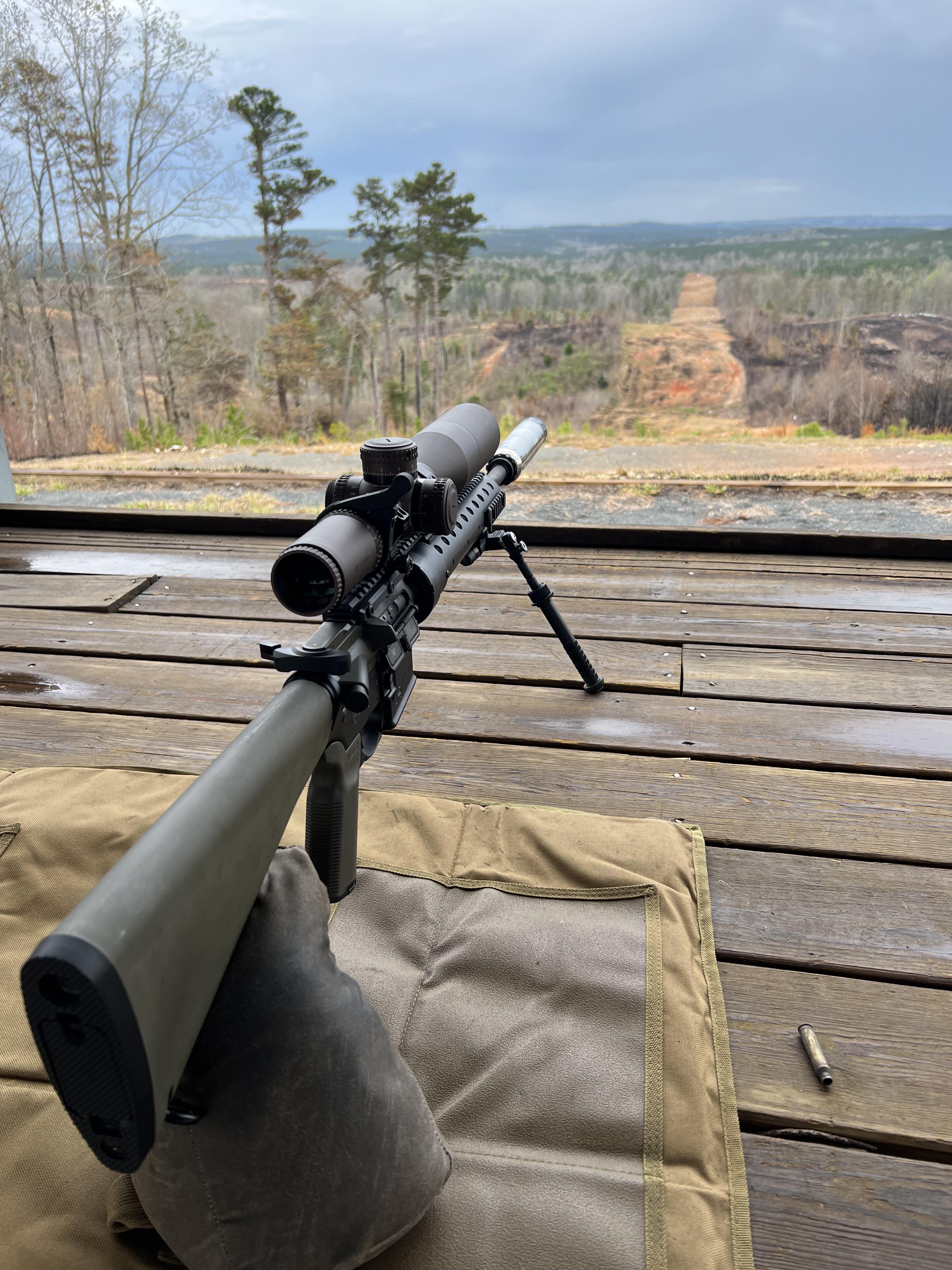 OD green AR-platform precision rifle with scope, suppressor, and bipod resting on shooting bags at an outdoor long-range bench, with a wooded valley downrange.