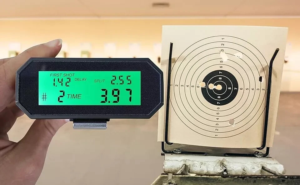 Hand holding a shot timer with green LCD display showing shot timing data next to a paper bullseye target with two bullet holes on a range target stand.