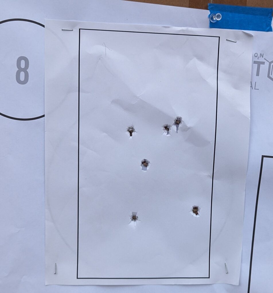Paper target with six bullet holes spread across the center area, stapled to a backer board at an outdoor range.