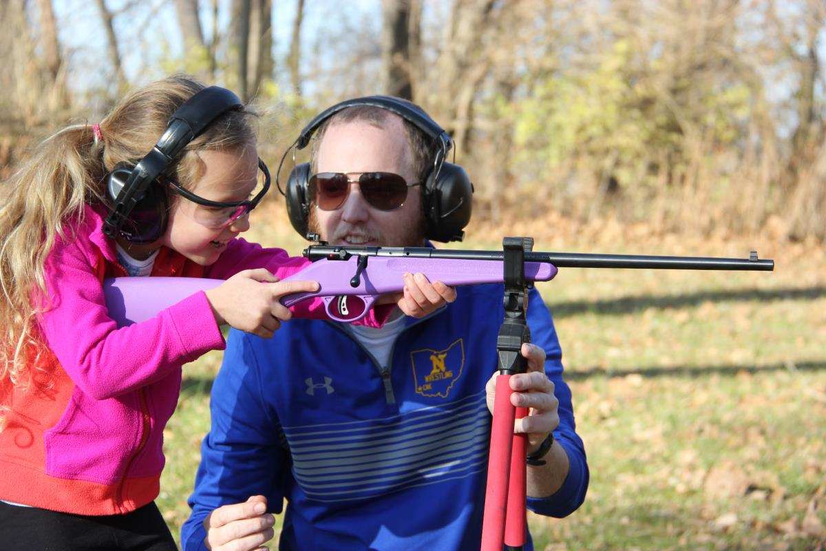 Adult helping a young girl aim a purple youth rifle outdoors, both wearing hearing protection and safety glasses.