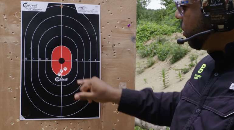 Person pointing to bullet holes on a Caldwell paper target at an outdoor shooting range.