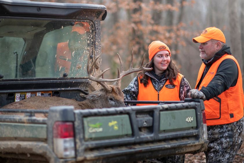 Young female hunter in blaze orange standing beside an older male hunter at the tailgate of a 4x4 utility vehicle with a harvested whitetail buck in the bed, in a wooded setting during deer season.