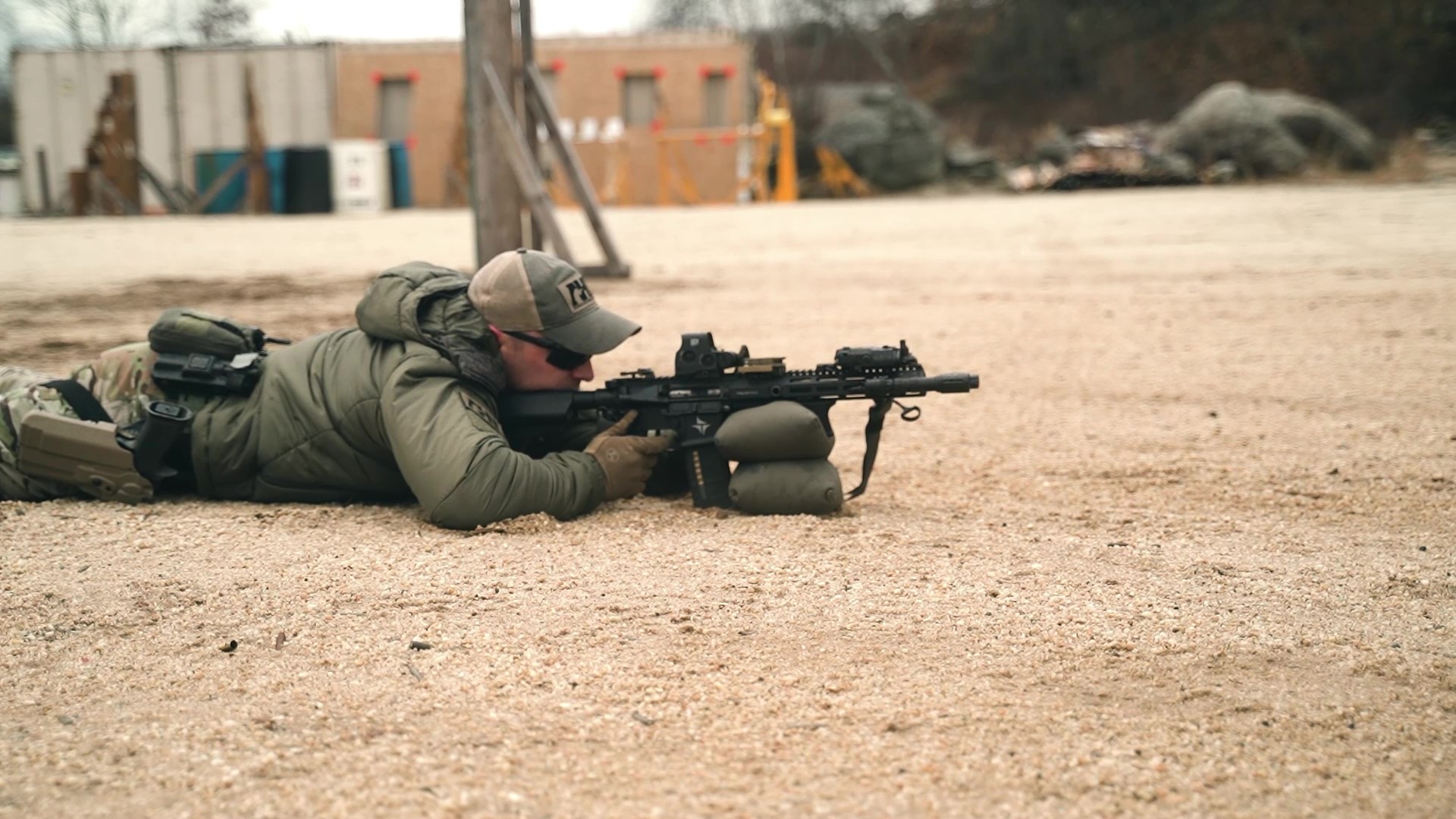 Shooter in prone position aiming an AR-platform rifle with red dot and magnifier on a gravel shooting range.