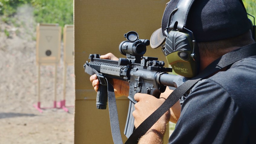 Shooter at outdoor range aiming an AR-style rifle with red dot optic and vertical foregrip, wearing electronic earmuffs