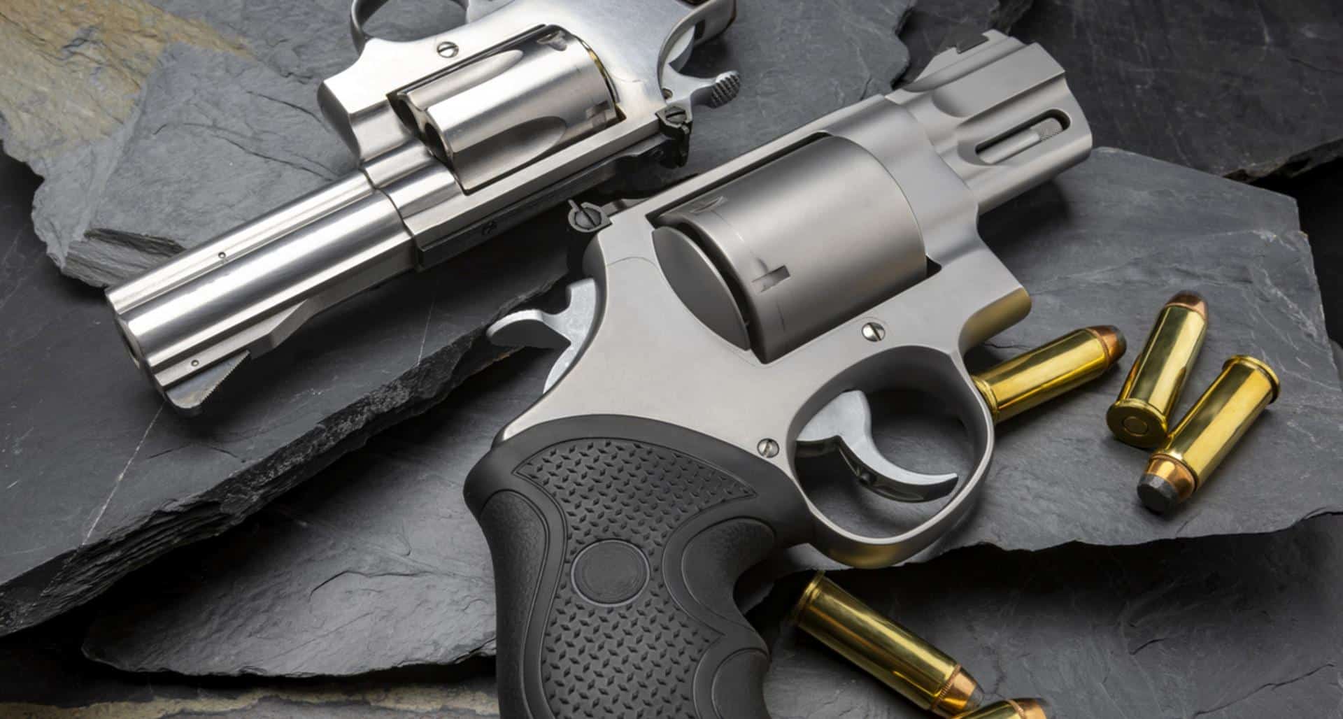 Two stainless revolvers with black rubber grips on slate rock surface with loose brass cartridges nearby.
