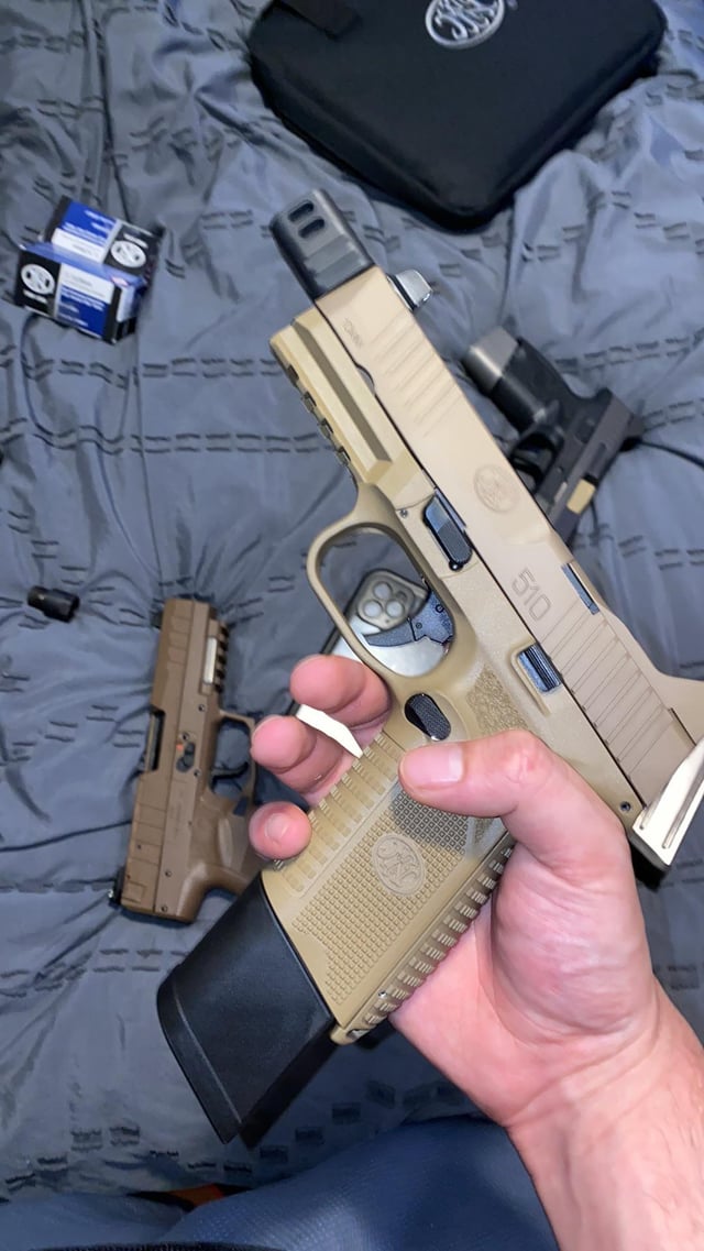Hand holding an FN 510 pistol in flat dark earth finish, showing grip texture and extended magazine, with additional firearms and ammo boxes on a gray surface behind it.