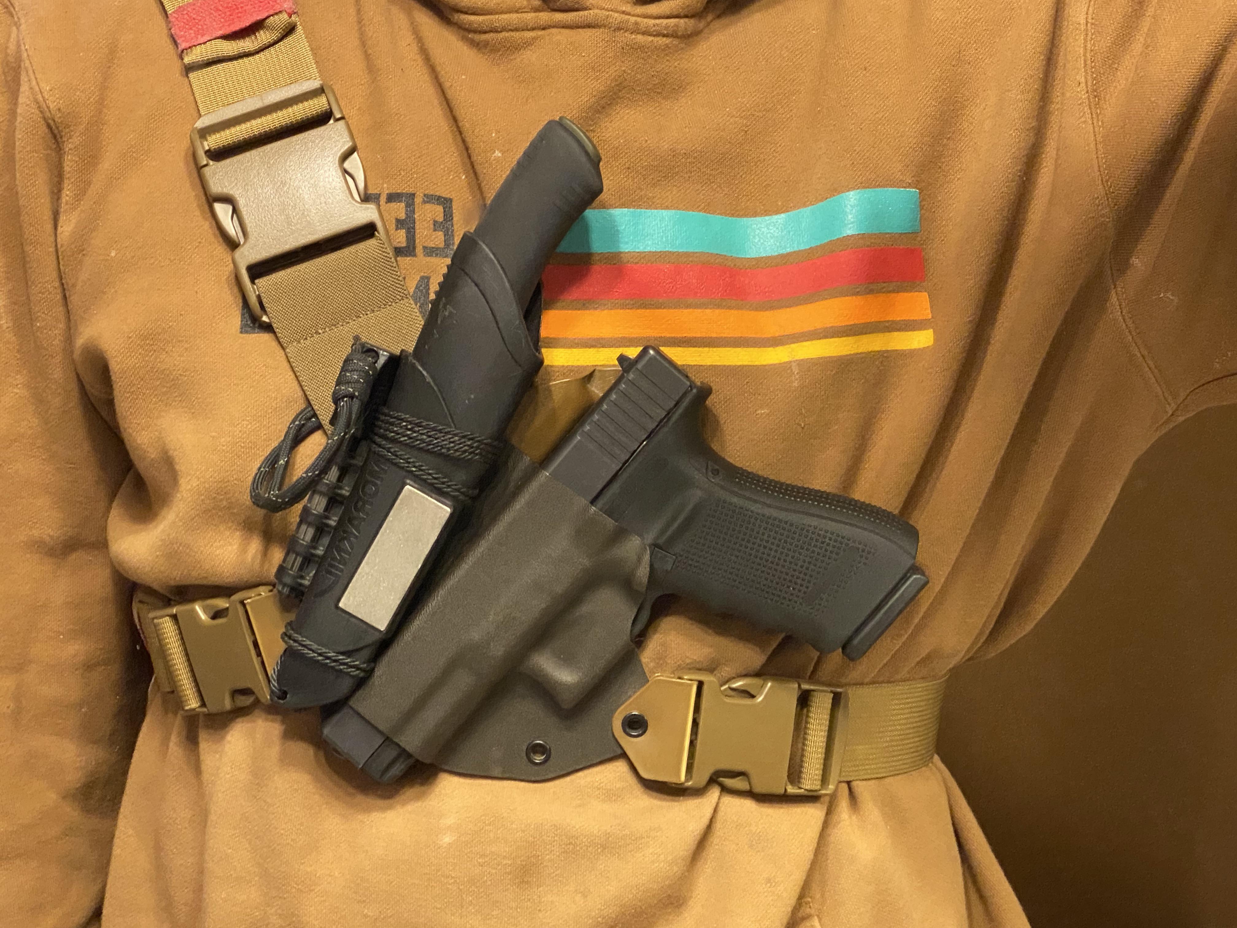Person wearing a chest holster carrying a Glock pistol, mounted on a coyote tan belt and shoulder harness system over a brown hoodie.