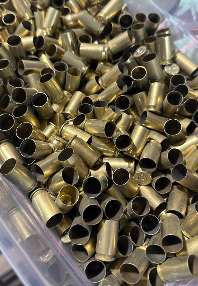 Large quantity of once-fired brass pistol casings piled in a clear plastic bin, viewed from above.