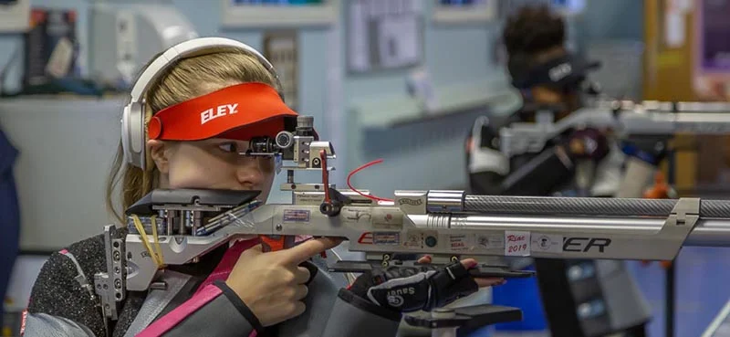 Female competitive shooter aiming a precision air rifle at an indoor range, wearing an ELEY visor and hearing protection.