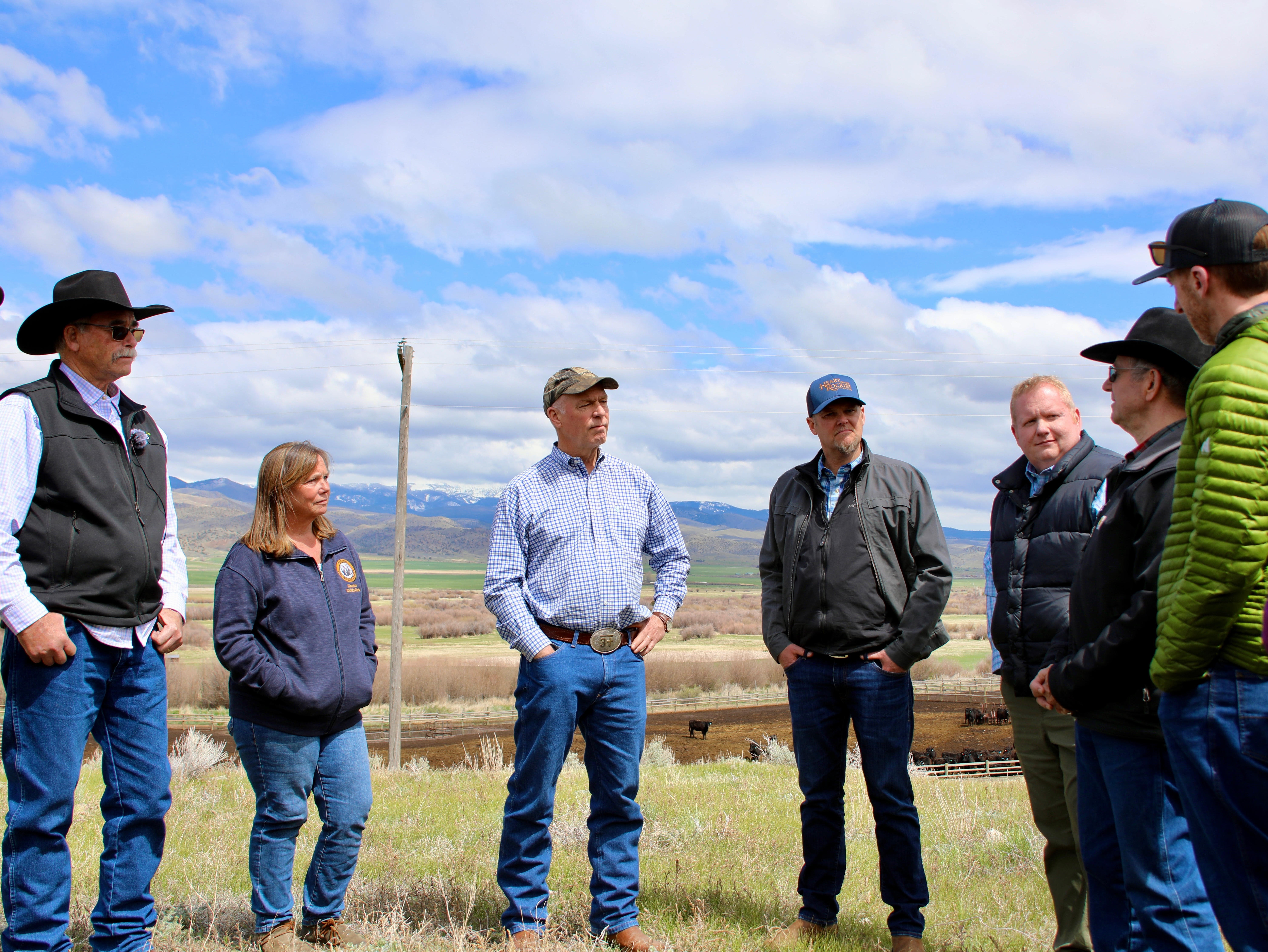 Group of seven people standing in a circle outdoors on a ranch with cattle pens, sagebrush, and snow-capped mountains in the background.