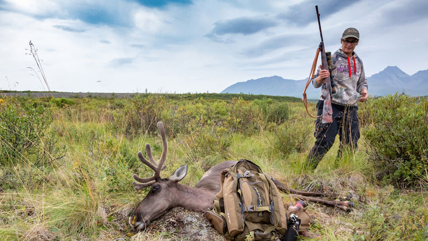 Hunter holding a bolt-action rifle standing next to a harvested caribou in an Alaskan tundra landscape with mountains in the background.