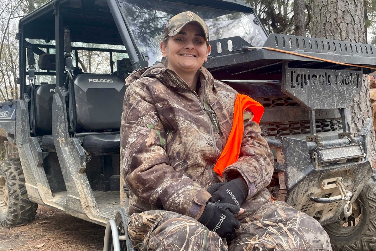 Woman in camouflage hunting gear and blaze orange vest seated in a wheelchair in front of a muddy Polaris UTV in a wooded area.