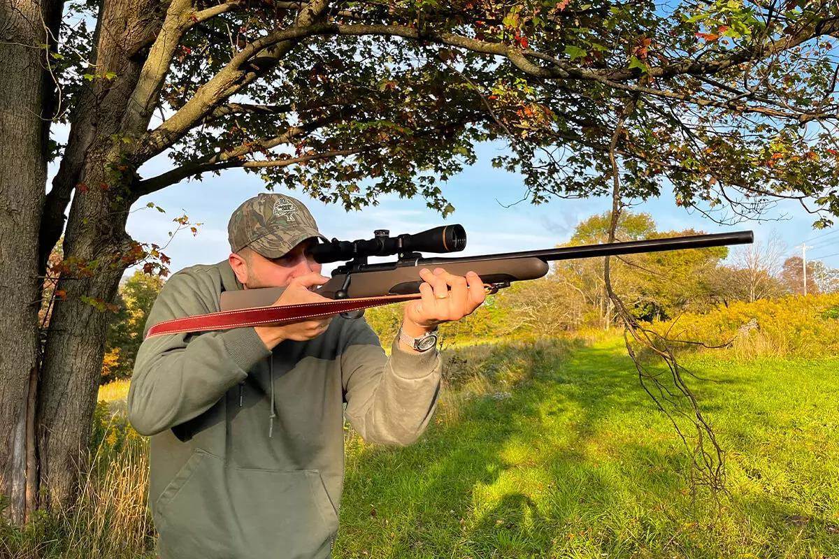 Man shouldering a scoped bolt-action rifle outdoors under a tree in autumn, aiming through the scope with a red leather sling visible.