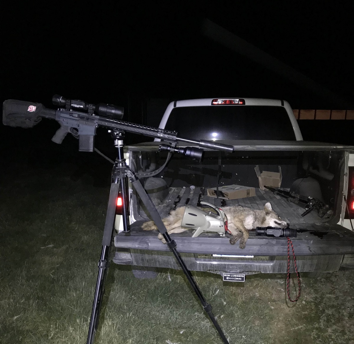 AR-style rifle with ATN night vision scope and suppressor on a tripod next to a truck bed containing a harvested coyote, electronic call, and thermal device at night.