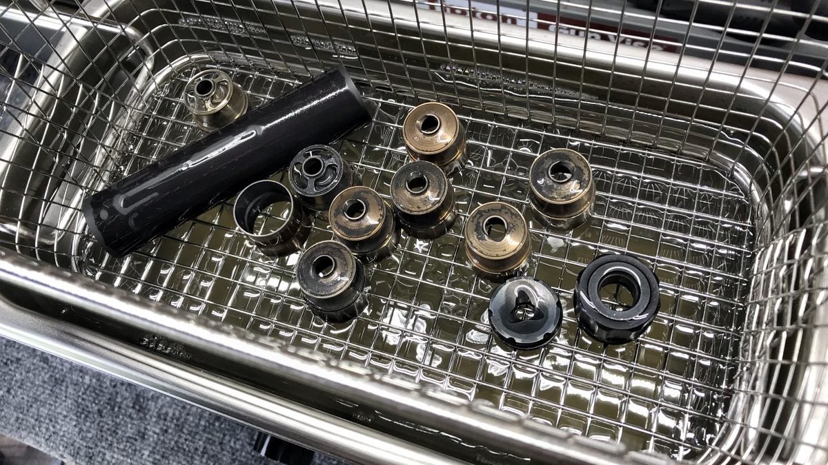 Disassembled rimfire suppressor baffles and a tube section sitting in a stainless steel ultrasonic cleaner basket, showing significant carbon fouling and lead deposits.