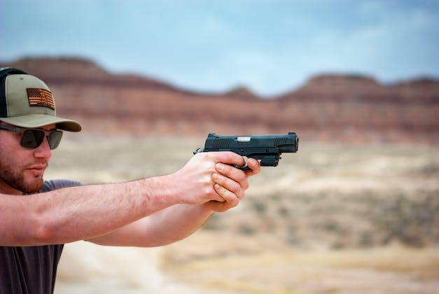 Man in baseball cap and sunglasses shooting a black semi-automatic pistol at an outdoor desert range, arms extended in a two-handed grip.